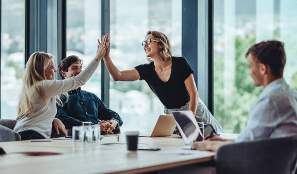 A25 Female professional giving a high five to her colleague in conference room. Group of colleagues celebrating success in a meeting.